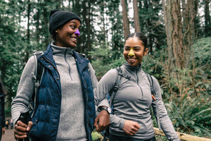 Two black women hiking with arms interlocked, with smiles on their faces, and bright Nöz sunscreen applied on their nose.  The woman  on the right is wearing the colorful Grape shade, and the woman on the left is wearing the fun Mellow Yellow shade. 